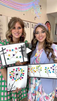 two beautiful women holding up paintings in front of a wall with crosses and flowers on them