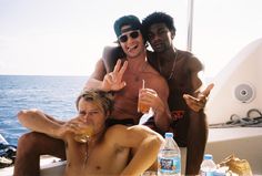 two men and a woman sitting on a boat with drinks in front of them, making the peace sign