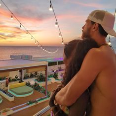 a man and woman standing on top of a balcony next to the ocean at sunset