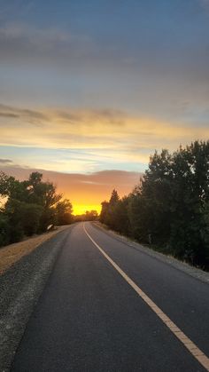 the sun is setting on an empty road with trees in the foreground and clouds in the background