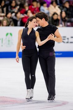 a man and woman in black ice skating on an ice rink with people watching from the stands