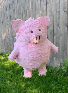 a pig made out of pink tissue paper sitting in the grass next to a fence