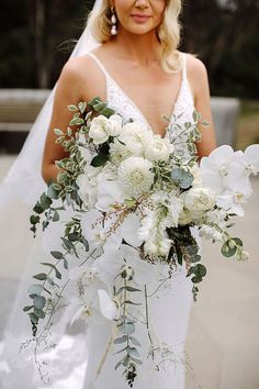 a woman in a wedding dress holding a bridal bouquet with white flowers and greenery