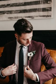 a man in a suit and tie adjusts his boutonniere while sitting on a couch