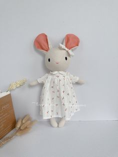 a white stuffed animal with pink ears and dress next to a book on a table