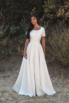 a woman wearing a white dress standing in front of some bushes and trees with her hands on her hips