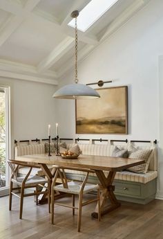 a dining room table and bench with two lights hanging from it's ceiling, in front of a painting on the wall