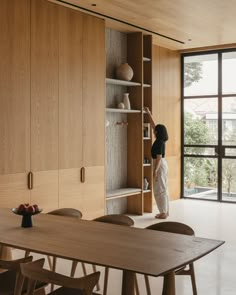 a woman standing in front of a wooden table next to bookshelves and shelves