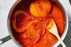 a pot filled with red sauce and onions on top of a white countertop next to a wooden spoon