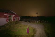 a woman in a red dress standing on a dirt road next to a barn at night