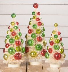 three small white christmas trees decorated with multicolored ornaments on top of a wooden table