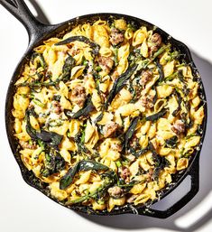 a skillet filled with pasta and vegetables on top of a white table next to a spoon