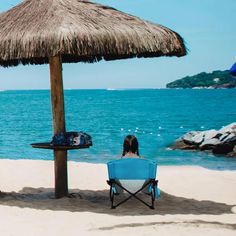 two people sitting in lawn chairs under an umbrella on the beach, facing the ocean