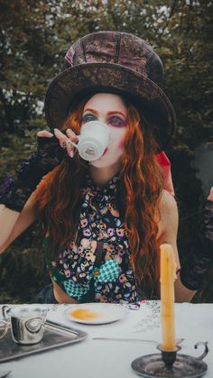 a woman with long red hair wearing a hat and drinking from a cup while sitting at a table