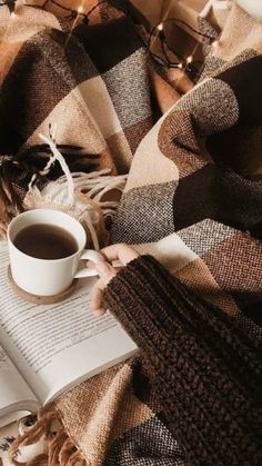 a person is reading a book and holding a cup of coffee while laying on a plaid blanket