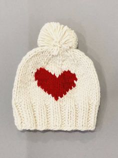 a white knitted hat with a red heart on the front and bottom, sitting on top of a gray surface