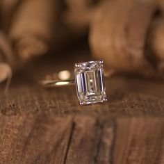 an emerald cut diamond ring sitting on top of a piece of wood with other items in the background