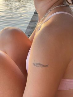 a woman with a fish tattoo on her arm sitting next to the water and looking off into the distance