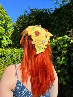 a woman with red hair wearing a yellow crochet sunflower hat in front of some bushes