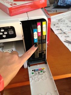 a person pointing at the ink cartridges in a printer's compartment on a desk