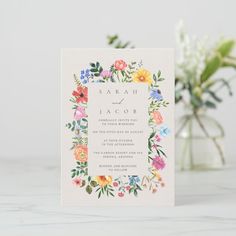 a wedding card with colorful flowers and greenery on the front, sitting next to a vase