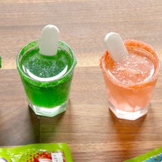 three different colored drinks sitting on top of a wooden table next to packets of gummy bears