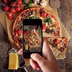 a person taking a photo of a pizza on their cell phone with cheese, tomatoes and mushrooms