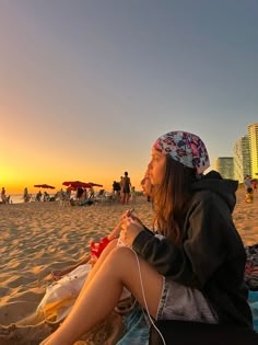 a woman sitting on top of a sandy beach next to the ocean with headphones in her ears