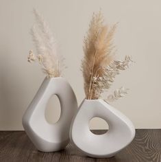 two white vases sitting on top of a wooden table next to each other with dried plants in them