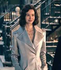 a woman standing in front of some stairs wearing a gray coat and smiling at the camera