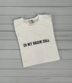 a white t - shirt with the words in my bride era printed on it, against a wooden background