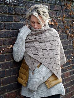 a woman leaning against a brick wall wearing a knitted shawl and jacket with pockets