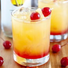 two glasses filled with drinks and cherries on top of a wooden table next to a bottle