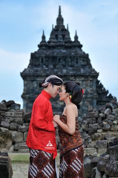 a man and woman standing next to each other in front of a stone structure with a sky background