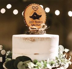 a wedding cake with a wooden slice that says just hooked on it and greenery around the edge