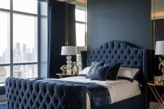 a bed with blue upholstered bedspread and pillows in front of large windows