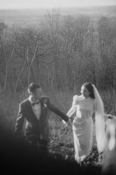 black and white photograph of a bride and groom walking through the woods with trees in the background