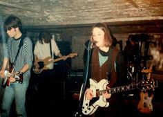 two young men are playing guitars and singing into microphones while another man stands behind them