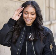 a woman with long hair wearing a black jacket and smiling at the camera while holding her hand up to her head