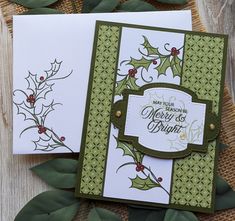 two cards with holly designs on them sitting next to some green leaves and greenery