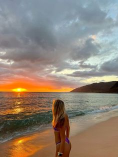 a woman standing on top of a sandy beach next to the ocean at sun set