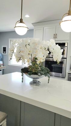 white flowers in a glass bowl on a kitchen countertop with lights above it and an oven