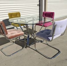 a glass table with four chairs around it on concrete ground next to wall and building