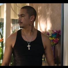 a man wearing a cross necklace and black tank top standing in front of a house