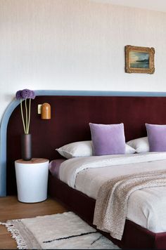 a bed with purple and white pillows in a bedroom next to a painting on the wall