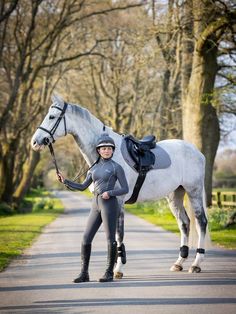 a woman standing next to a white horse in the middle of a road with trees