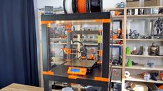 an orange and black 3d printer sitting on top of a wooden table next to shelves