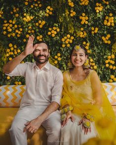 a man and woman sitting on a yellow couch in front of a wall with orange flowers