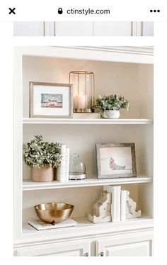 a white book shelf with some plants and pictures on it's shelves, next to a gold bowl