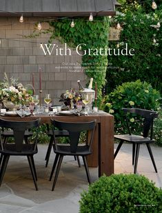 an outdoor dining table set for four with flowers and candles on it, surrounded by greenery